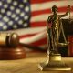 Scales of Justice statue in front of a American flag, a judges gavel, and books blurred in the background.