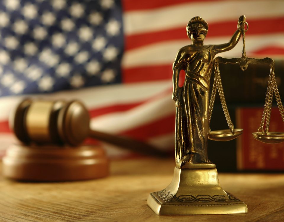 Scales of Justice statue in front of a American flag, a judges gavel, and books blurred in the background.