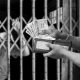 A person behind bars blurring off in the background with a person on the other side of the bars holding a wallet full of money.