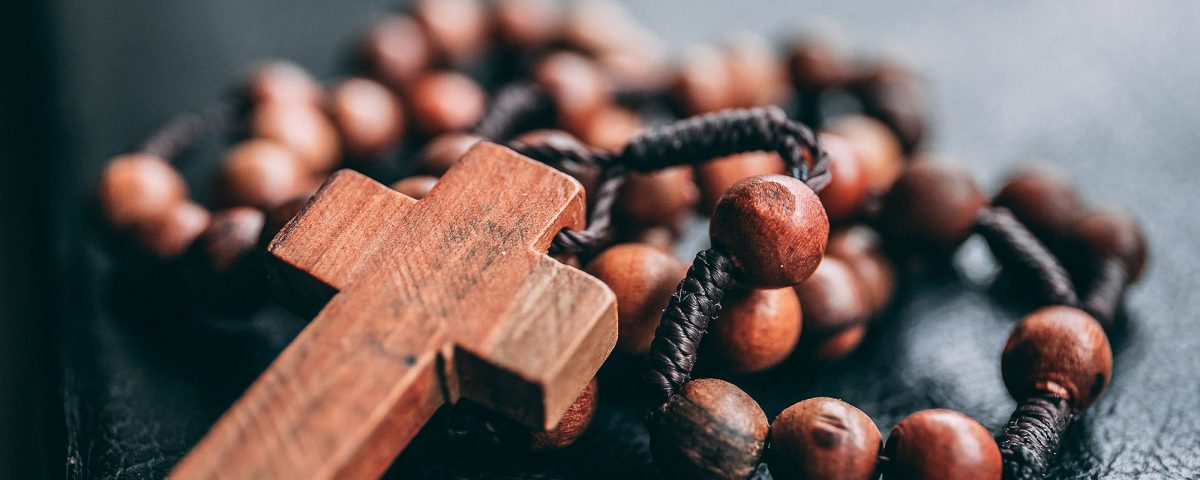 A photograph of rosary beads with the cross. The beads & cross are a red wood with the twine lacing them together. The twine is a deep brown that braids around the beads.