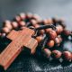 A photograph of rosary beads with the cross. The beads & cross are a red wood with the twine lacing them together. The twine is a deep brown that braids around the beads.