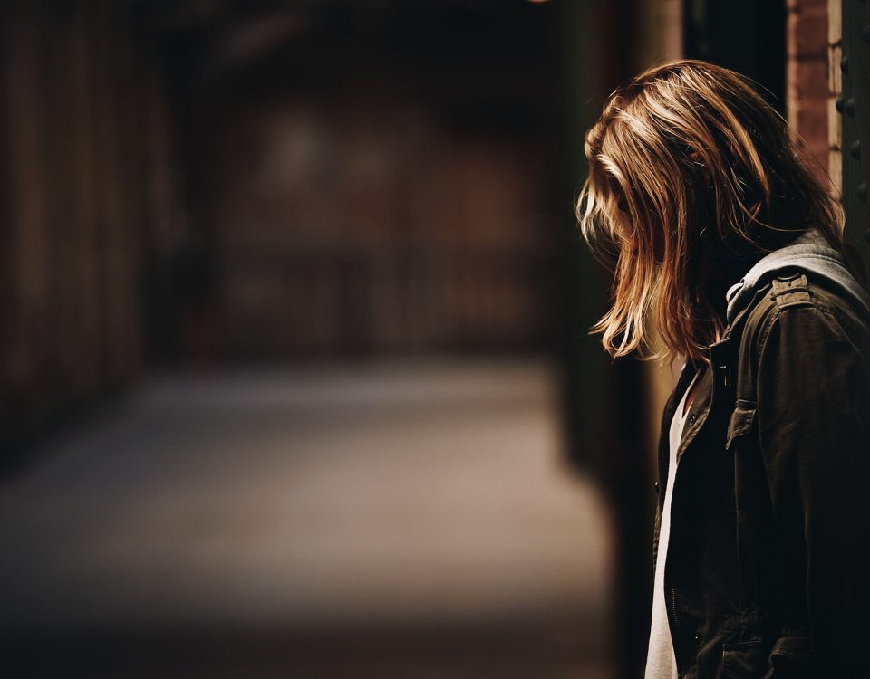 A photograph of a person with long hair covering their face. They're the focal point of the image, and the hallway they're in is blurred out. The person appears to be looking downward and seems sad or deep in thought.