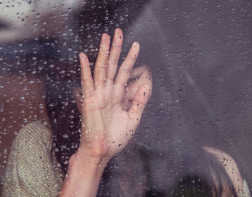 A person places their hand upon a glass window, the rain and condensation blurs the person out of view, but they appear to be sad or deep in thought.