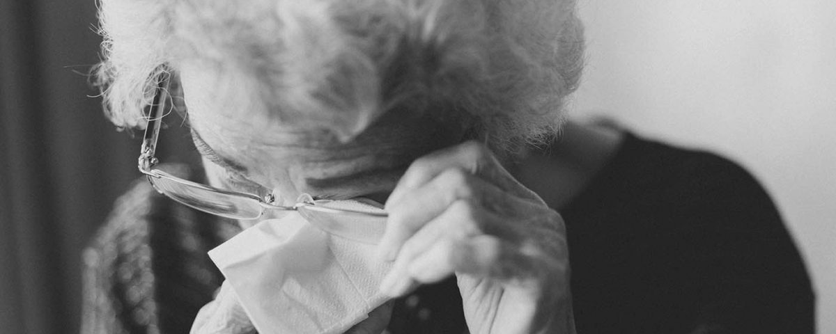 An elderly woman wiping away tears under her glasses, the image is in black and white and it seems that she is deeply sad.