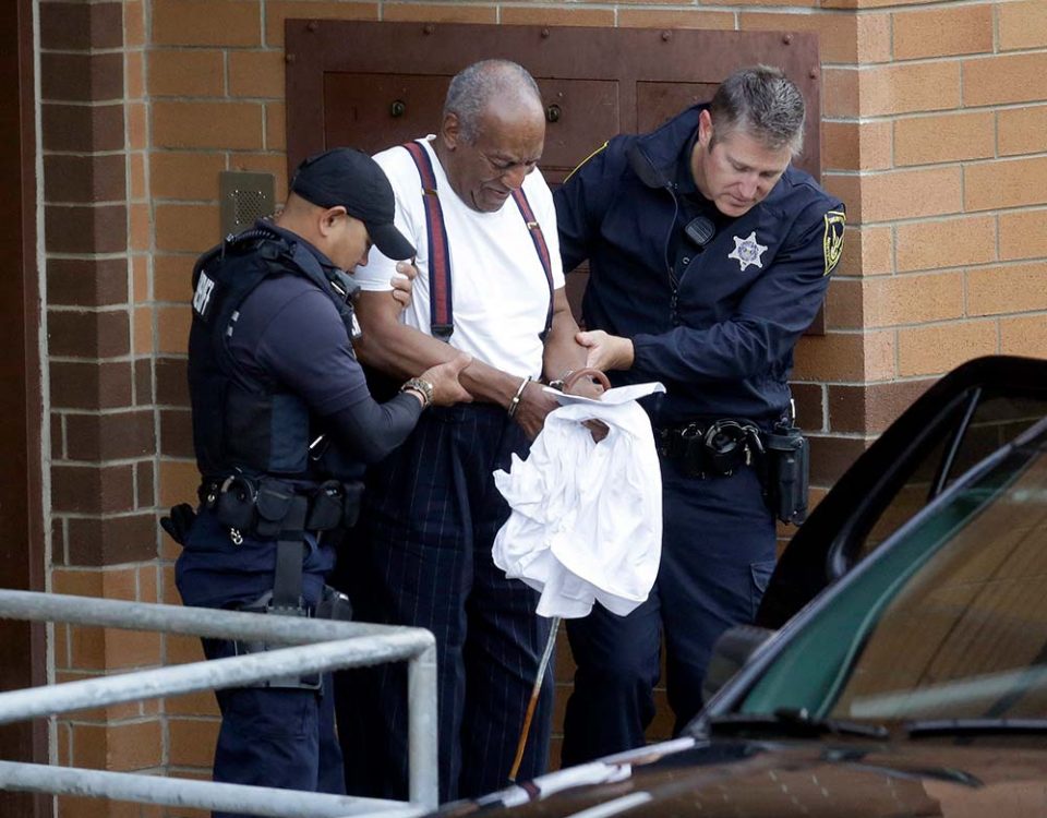 Bill Cosby, a famous comedian, is being escorted towards a vehicle in handcuffs by police officers.