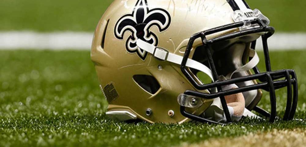 A football field is blurred out in the background, with a football helmet laid on the ground. The helmet is a "Saints" helmet which is a Louisiana team, and it has scuff marks from use on the helmet.