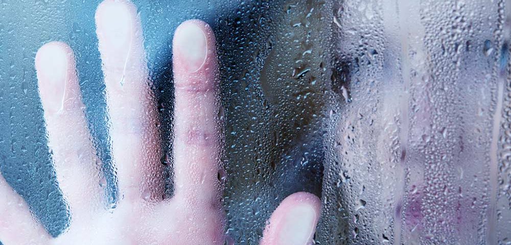 A person places their hand upon a glass window, the rain and condensation blurs the person out of view, but they appear to be sad or deep in thought.