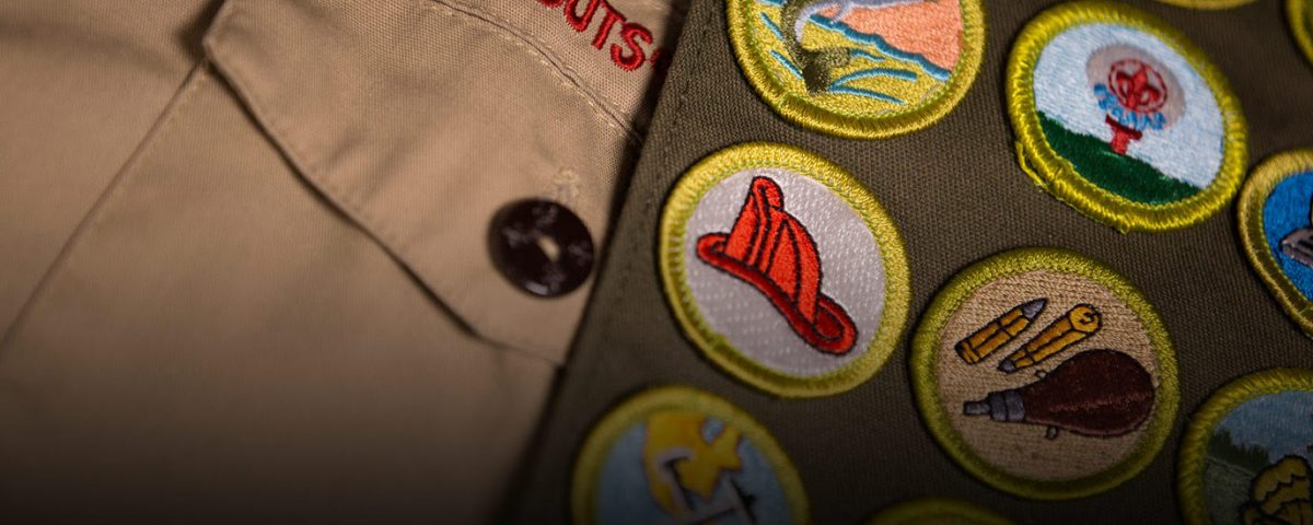 Close up of Boy Scout uniform, showing the badges sewn into the sash. The name Boy Scout can clearly be seen, with a fireman helmet badge as the most visible badge being displayed.
