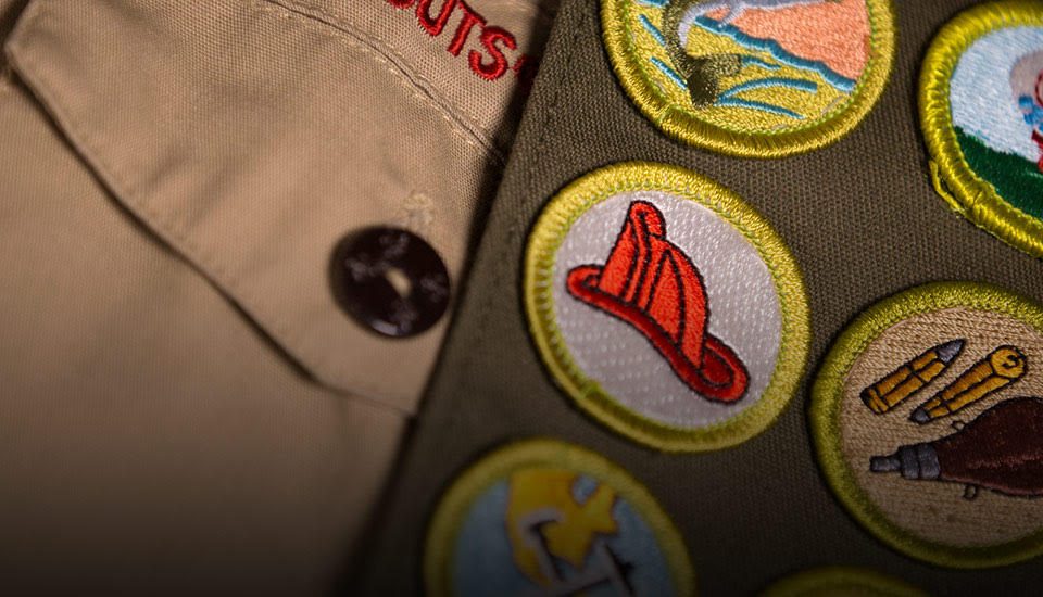 Close up of Boy Scout uniform, showing the badges sewn into the sash. The name Boy Scout can clearly be seen, with a fireman helmet badge as the most visible badge being displayed.